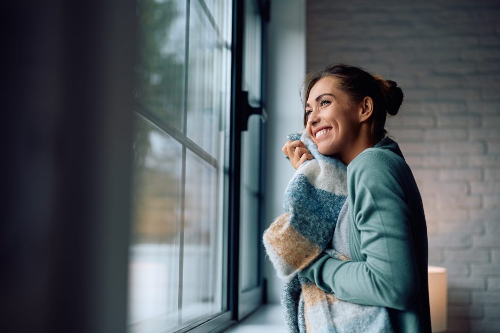 Home happy woman holding a blanket while standing by the window and l